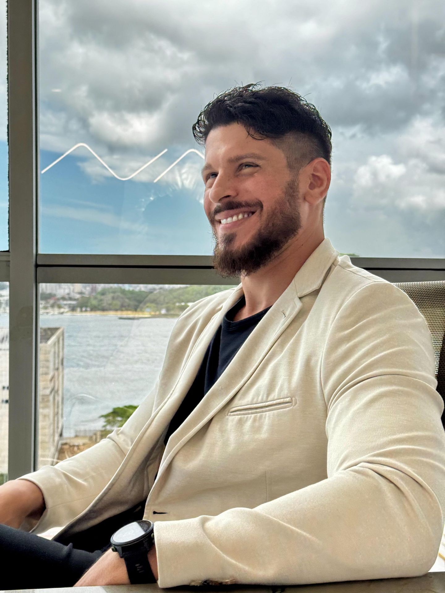 Man in a beige blazer sitting by a window with a view of water and cloudy sky.