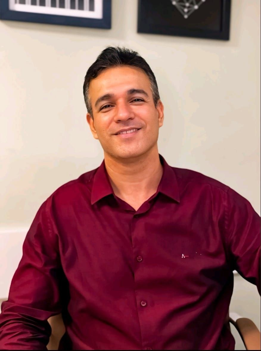 Smiling man wearing a red shirt, sitting indoors with framed artwork on the wall.