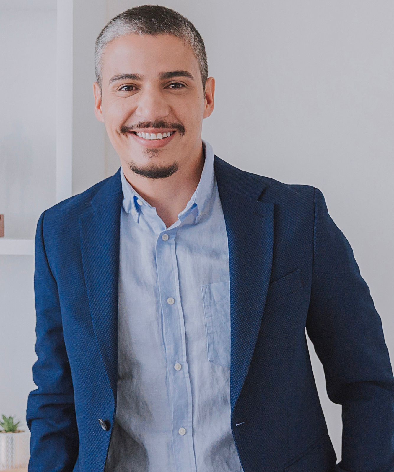 Smiling person in a blue blazer and light blue shirt standing indoors.