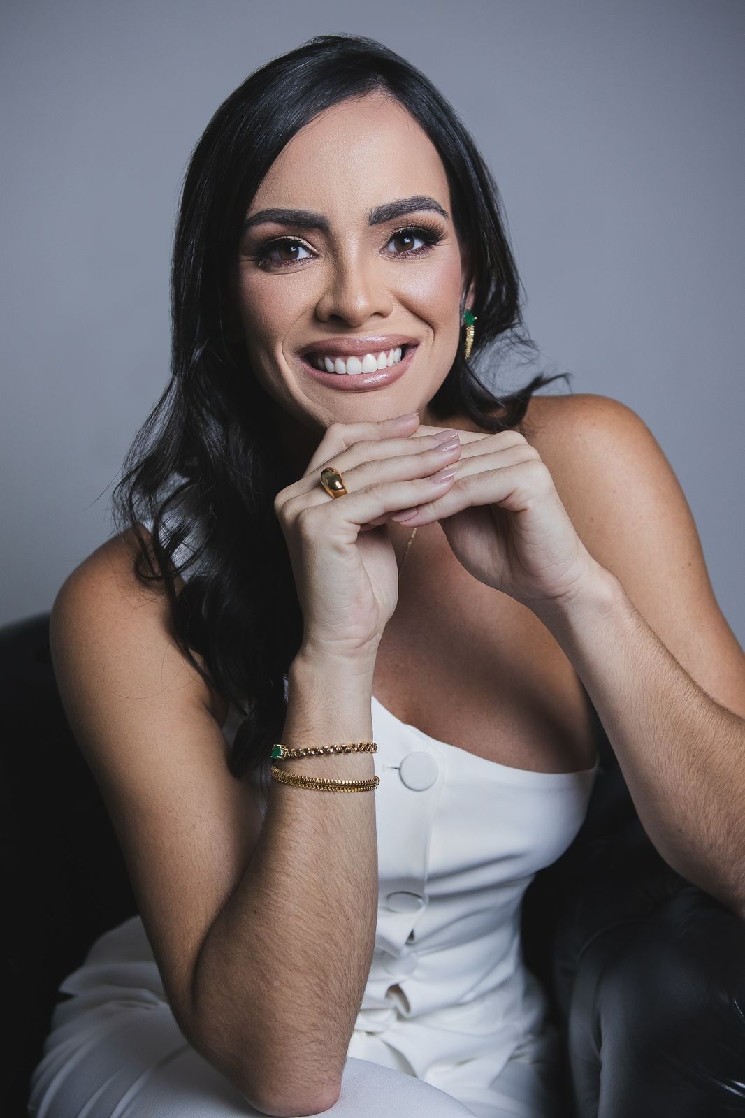 Smiling person with long dark hair and white sleeveless outfit, resting chin on hands.